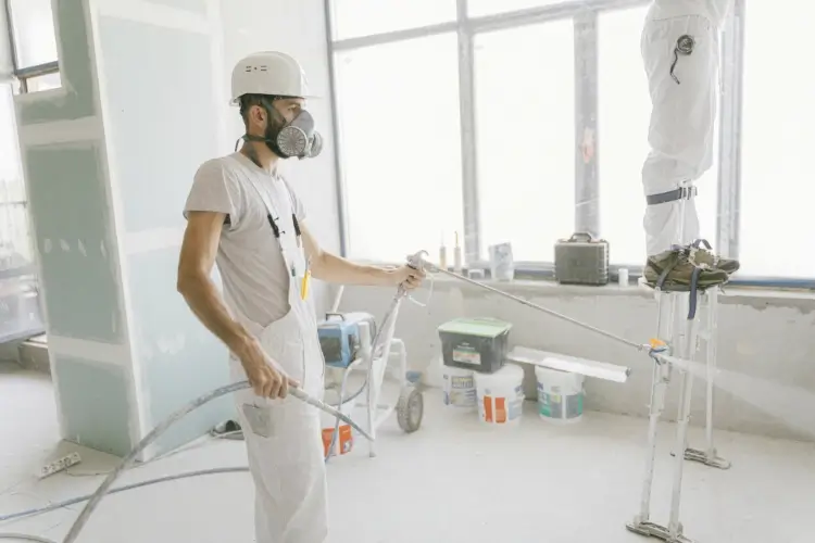Two construction workers indoors, one on stilts, wearing protective gear with construction materials around them.
